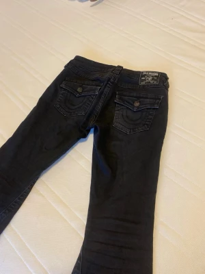 Svarta jeans från True Religion - Säljer ett par svarta jeans från True Religion med snygga ficklock och dekorativa sömmar på bakfickorna. Jeansen har klassisk femficksdesign, knappstängning och bälteshällor. Materialet är mjukt och stretchigt för skön passform.