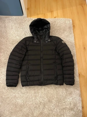 Svart Colmar dunjacka med huva - Snygg svart dunjacka från Colmar Originals med huva och två praktiska fickor med dragkedja. Jackan har ett quiltat mönster och logotyp på ärmen. Perfekt för kalla dagar och har en stilren look som funkar till det mesta.