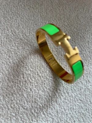 Grön och guld  Clic  armband - Säljer ett  Clic armband i guld med gröna paneler och den ikoniska -detaljen framtill. Armbandet har en stilren och modern design med blank finish och är tillverkat i metall med emalj. Perfekt accessoar för att lyfta din outfit.