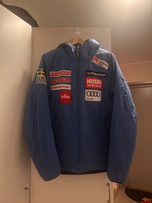 Blå vindjacka Huski Wear Team Sweden - Blå vindjacka från Huski Wear med Team Sweden Alpine-tryck och flera sponsormärken som Bauhaus, Audi och Skistar. Jackan har huva med gul snörning, hel dragkedja och svart quiltad insida. Perfekt för dig som gillar sportig stil och alpina vibes. Perfekt för skidåkning.