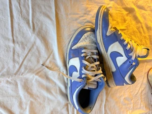 Nike Dunk Low blå/vita sneakers - Säljer ett par Nike Dunk Low sneakers i blått och vitt skinn med klassisk Nike Swoosh på sidorna. Skorna har vit snörning, rund tå och platt sula. Yttersulan är blå och hälen har broderad Nike-logga. Perfekta för dig som gillar retrovibbar och streetwear. Dessa är välanvända men har kvar att ge för den som orkar putsa dom lite.