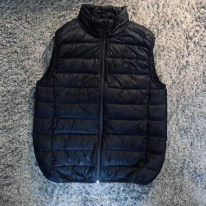 Svart dunväst från Lindbergh XL - Snygg svart dunväst från Lindbergh i storlek XL. Västen har quiltad design, hög krage och dragkedja framtill. Tillverkad av återvunna syntetfibrer och har en diskret rund patch på bröstet. Perfekt för lager på lager och chill stil.