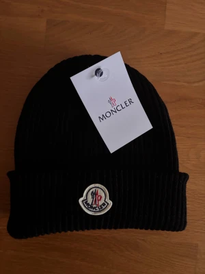 Svart ribbad mössa från Moncler - Säljer en svart ribbstickad Moncler-mössa med klassisk Moncler-logga framtill. Mössan är i mjukt material och har en uppvikt kant för extra stilpoäng. Perfekt accessoar för kalla dagar och snygg till alla outfits. Skickar snabbt:)