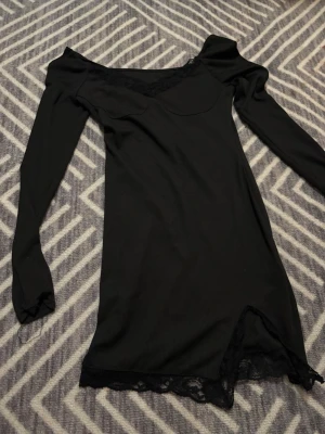 Svart långärmad klänning med spets - Svart ribbad klänning med långa ärmar och offshoulder-skärning. Klänningen har v-ringad hals med spetsdetaljer och spetskant nertill samt en liten slits framtill. Snygg och enkel modell. Märke står ej eller storlek, men passar mig som är S.