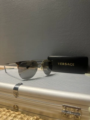 Versace solglasögon guld och svart - Säljer ett par riktigt snygga Versace solglasögon med guldfärgade metallbågar och svarta skalmar. Glasen är tonade i grått och har en klassisk pilotform. Medföljer originaletui med Versace-logga. Perfekt accessoar för dig som vill sticka ut.