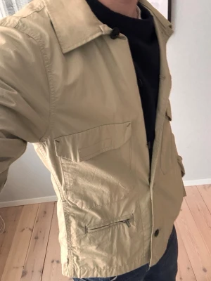 Beige overshirt från G-Star RAW - Snygg overshirt från G-Star RAW med klassisk krage, stora fickor framtill och knäppning med svarta knappar. Jackan har lång ärm och en cool zip-detalj på ena fickan. Perfekt att slänga på över en hoodie eller t-shirt. Storlek S! 
