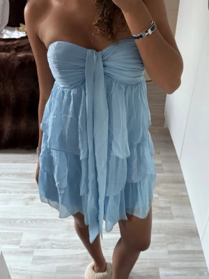 Ljusblå axelbandslös volangklänning Nelly  - Supersöt ljusblå kortklänning med flera lager volanger och en stor knytning framtill. Klänningen är från Nelly och andvänd 1 gång, sitter som en xs/s