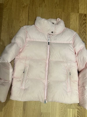 Ljusrosa pufferjacka från Monel XS - Säljer en ljusrosa pufferjacka från Monel i storlek XS. Jackan har hög krage, två sidofickor med dragkedja och en snygg Monel-logga på ärmen. Materialet är mjukt och syntetiskt, perfekt för kalla dagar. Dragkedja framtill och quiltad design för extra värme.