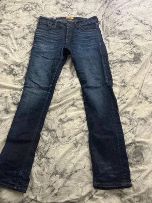 Jack & Jones Slim Fit Tim Jeans - Mörkblå jeans från Jack & Jones i modellen Slim Fit Tim. Klassisk femficksdesign, snygga slitningar och smal passform. Tillverkade i mjukt denimtyg med subtila kontrastsömmar och läderpatch bak i midjan.