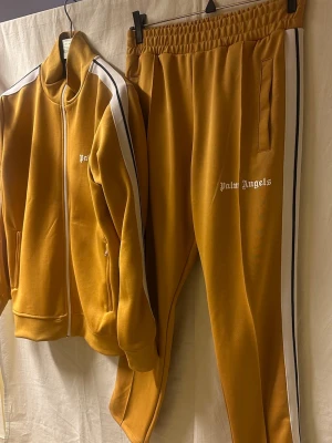 Mustardgul Palm Angels tracksuit - Palm Angels tracksuit i en snygg mustardgul färg med vita och svarta ränder längs sidorna. Aldrig använd endast testad. 