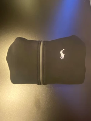 Svart hoodie från Polo Ralph Lauren - Svart hoodie från Polo Ralph Lauren med klassisk vit broderad logga på bröstet. Tröjan har huva, hel dragkedja och två fickor framtill. Insidan är mjuk och fleecefodrad för extra komfort. Perfekt för chill dagar och streetwear-stil.