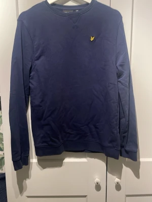 Mörkblå sweatshirt från Lyle & Scott - Mörkblå sweatshirt från Lyle & Scott med klassisk rund hals och gul broderad logga på bröstet. Tröjan har långa ärmar, ribbade muddar och är tillverkad i mjuk bomull. Perfekt för en chill och stilren look. Stollen är ungefär en XS. 