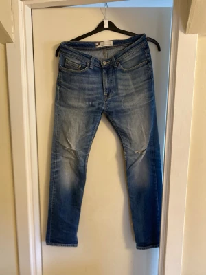 Blå jeans med slitningar på knäna - Snygga blå jeans med klassisk femficksdesign och slitna detaljer på båda knäna. Jeansen har en rak passform och är tillverkade i jeansmaterial med lätt tvättad look. Perfekta för dig som gillar en avslappnad och cool stil.