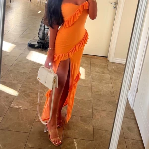 Orange långklänning med volanger - Säljer en snygg orange axelbandslös långklänning med volangdetaljer och hög slits framtill. Klänningen har en figurnära passform och är perfekt för dig som vill sticka ut. Materialet känns mjukt och följsamt mot kroppen. Perfekt för sommarens alla tillfällen!