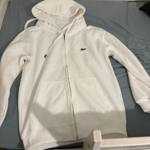 Vit hoodie från Lacoste med dragkedja - Vit hoodie från Lacoste med klassisk grön krokodillogga på bröstet. Tröjan har huva med snörning, dragkedja framtill och två stora fickor. Perfekt för en chill och stilren look. Helt oanvänd!