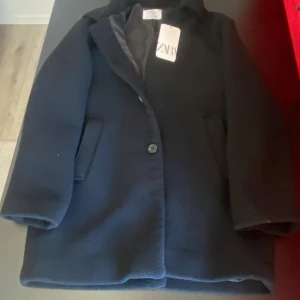 Svart kappa från Zara, stl 164 - Stilren svart kappa från Zara i klassisk rak modell med krage och knäppning framtill. Jackan har två fickor på sidorna och är helfodrad för extra komfort. Perfekt för dig som gillar en clean och tidlös look. Stl 164 men passar S