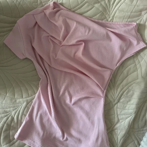 Ljusrosa ribbad offshoulder topp - Säljer en ljusrosa ribbad topp med offshoulder-design. Toppen har en tight passform och är tillverkad i ett mjukt, stretchigt material som sitter snyggt på kroppen. Perfekt för dig som gillar en enkel men trendig stil.