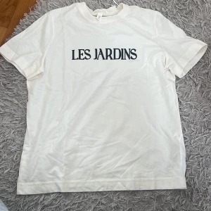 Vit t-shirt med tryck LES JARDINS - Snygg vit t-shirt med svart texten 'LES JARDINS' på bröstet. Klassisk passform med rund hals och korta ärmar. Perfekt till jeans eller shorts för en clean och stilren look.