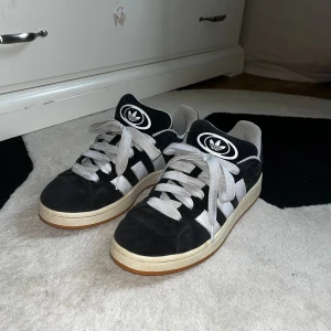 Adidas Campus 00s svart - Svarta Adidas Campus 00s skor i mocka med vita detaljer. Skorna har beige sula, vit snörning och stor Adidas-logga på plösen. Perfekt för dig som gillar retrostil och streetwear.