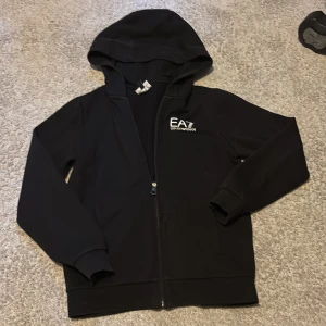 Svart EA7 hoodie med dragkedja - Svart hoodie från EA7 Emporio Armani med vit logga på bröstet. Tröjan har huva, långa ärmar och hel dragkedja framtill. Mjuk insida och ribbade muddar vid ärmslut och nederkant. Perfekt för en chill och stilren look. Nypris ca 1100kr pris är inte hugget i sten.