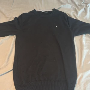 Svart stickad tröja från J.Lindeberg - En stilren svart stickad tröja från J.Lindeberg med diskret logga broderad på bröstet. Tröjan har rund halsringning, ribbade muddar och är långärmad. Perfekt för dig som gillar klassisk och enkel design.