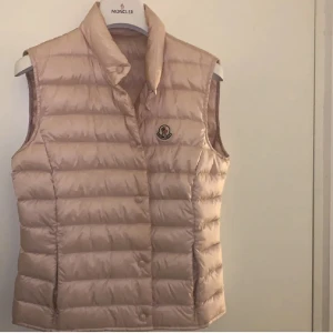 Moncler väst - Snygg rosa dunväst från Moncler. Västen har tryckknappar framtill och ikonisk Moncler-logga på bröstet. Lite smink vid kragen men det ska gå bort i tvätten! Dustbag och moncler taggarna på sista bilden medföljer!