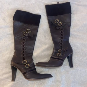Bruna höga stövlar med snörning och klack - Snygga bruna stövlar från Marco Tozzi i skinn med högt skaft, spetsig tå och hög klack. Dekorativa snörningar och spännen i mässing samt dragkedja på insidan. Ovankant i mörkare material som ger en cool kontrast. Perfekta för dig som vill sticka ut.