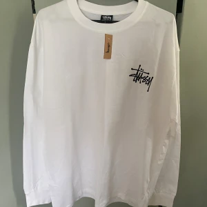 Vit långärmad Stüssy tröja XL - Vit långärmad tröja från Stüssy i bomull med svart logga tryckt på bröstet och stort tryck på ryggen. Klassisk rund halsringning och relaxed passform, perfekt för en streetwear-look.