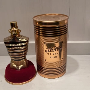 Jean Paul Gaultier Le Male Elixir - Exklusiv parfym i guldig metallflaska formad som en manlig torso, med matchande guldig burk och röd sammetsbas. Le Male Elixir är en ikonisk doft med lyxig och modern design, perfekt för dig som vill sticka ut.