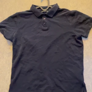 Svart pikétröja i bomull - En klassisk mörk blå pikétröja med krage och tre knappar framtill. T-shirten är kortärmad och tillverkad i mjuk bomull, perfekt för en clean och enkel stil. Passar dig som gillar basic och tidlösa plagg.