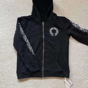 Svart hoodie från Chrome Hearts - Svart hoodie från Chrome Hearts med vita broderade detaljer längs ärmarna och logga på bröstet. Tröjan har huva, dragkedja framtill och två fickor. Perfekt för dig som gillar streetwear och snygga detaljer.