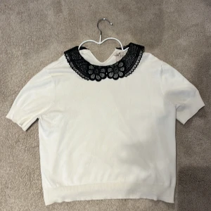 Vit topp med svart spetskrage H&M Divided - Vit topp från H&M Divided med korta ärmar och en snygg svart spetskrage med blommönster. Mjuk och skön känsla, perfekt för dig som gillar detaljer. Kragen ger en cool kontrast och gör toppen unik. Har krympt lite i tvätten.
