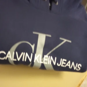 Blå hoodie från Calvin Klein Jeans - Säljer en snygg marinblå hoodie från Calvin Klein Jeans i storlek XS. Tröjan har stor vit CK-logga och text framtill samt snörning i huvan. Perfekt för dig som gillar stilrena och bekväma plagg med ikonisk branding. Finns att hämta i Grums tar bara cash
