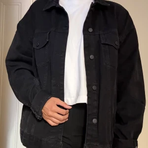 Svart oversized jeansjacka - Säljer en svart oversized jeansjacka med klassisk krage och knäppning framtill. Jackan har två bröstfickor med lock och knappar samt långa ärmar med knapp vid ärmslut. Perfekt att slänga över en hoodie eller t-shirt för en chill look.