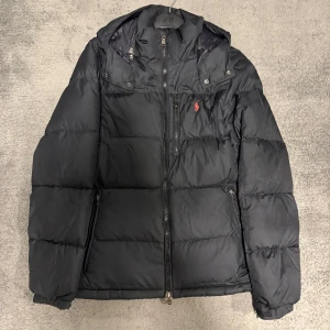 Svart dunjacka Polo Ralph Lauren - Svart dunjacka från Polo Ralph Lauren med röd broderad logga på bröstet. Jackan har huva, dragkedja framtill och två sidofickor med dragkedja. Puffig modell som håller dig varm under kalla dagar. Klassisk och stilren design. Perfekt inför vintern!🤩