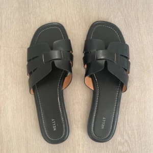Svarta sandaler från Nelly - Stilrena svarta sandaler från Nelly med fyrkantig tå och breda remmar i skinnimitation. Snyggt flätad design över foten och platt sula, perfekta för sommardagar. Enkla att matcha till olika outfits.