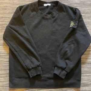 Svart sweatshirt från Stone Island - Svart sweatshirt från Stone Island med klassisk rund hals och ribbade muddar. Ikonisk logotyp-patch på vänster ärm med gula och gröna detaljer. Mjuk insida och stilren design. Om ni har några frågor så är de bara att höra av er! 