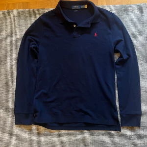 Polo Ralph lauren tröja  - Snygg marinblå långärmad pikétröja från Polo Ralph Lauren i slim fit. Tröjan har klassisk krage, knappar vid halsen och den ikoniska röda logon broderad på bröstet. Tillverkad i mjuk bomull som är skön mot huden. Perfekt för en clean och stilren look.