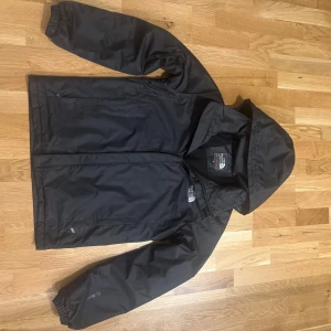 Svart vindjacka från The North Face M - Svart vindjacka från The North Face Summit Series med huva och dragkedja framtill. Jackan har broderad logga på bröstet och insidan, samt två sidofickor med dragkedja. Materialet är lätt och syntetiskt, perfekt för blåsiga dagar. Snygg och funktionell design.