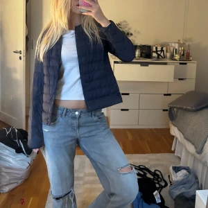 Mörkblå quiltad Uniqlo jacka - Säljer en mörkblå pufferjacka med quiltad design och raka linjer. Jackan har en enkel, stilren look utan krage och stängs med knappar framtill. Perfekt för dig som gillar en clean och chill stil. Passar till jeans och sneakers.