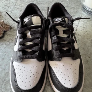 Nike Dunk Low svart/vit sneakers - Nike Dunk Low sneakers i svart och vitt med klassisk Swoosh på sidorna. Skorna har snörning, perforerad tå och platt sula. Ovandelen är i skinn och syntet, vilket ger en clean och tidlös look. Perfekta för dig som gillar streetstyle och vill ha en ikonisk sneaker.