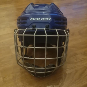 Blå hockeyhjälm Bauer med galler - Säljer en blå hockeyhjälm från Bauer med silverfärgat galler för ansiktsskydd. Hjälmen har ventiler på ovansidan och sidorna, justerbar rem och tydlig Bauer-logga på flera ställen. Perfekt för dig som spelar hockey och vill ha bra skydd på isen.