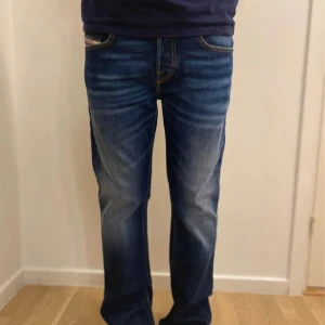 Blå jeans från Diesel med raka ben - Säljer ett par klassiska blå jeans från Diesel med snygga slitningar och kontrastsömmar. Jeansen har raka ben, normal passform och fem fickor. Märkesdetalj på myntfickan och bakfickor med dekorativa sömmar. Perfekta till sneakers eller boots.