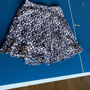 Blommig volangkjol från Seaside - Supersöt kort kjol från Seaside med mörkblå botten och vita blommor med röda detaljer. Kjolen har volang nertill och resår i midjan för skön passform. Perfekt för dig som gillar en lekfull och somrig stil!