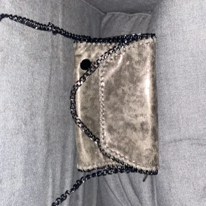 Glittrig silver axelväska med kedja - Snygg axelväska i glittrigt silver med svart kedjerem och dekorativa sömmar längs kanterna. Väskan har lockstängning och ett stort fack med dragkedja inuti. Perfekt för dig som vill sticka ut med en cool accessoar.