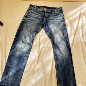 Blå jeans från Jack & Jones, 34/32 - Snygga blå jeans från Jack & Jones med klassisk femficksdesign och ljusa slitningar framtill. Jeansen har normal passform och raka ben, tillverkade i slitstarkt denim med orangea sömmar och läderpatch bak i midjan.