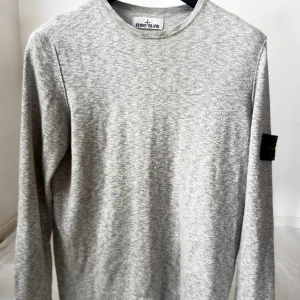 Grå långärmad tröja från Stone Island - Snygg grå långärmad tröja från Stone Island med klassisk rund hals och ikonisk logotyp-patch på vänster ärm. Tröjan är i mjuk bomull och har en stilren, enkel design som passar perfekt till jeans eller joggers.