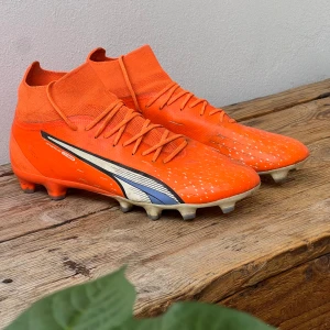 Orange fotbollsskor från Puma - Snygga orangea fotbollsskor från Puma med högt skaft och snörning. Skorna har vit och blå detalj på sidan och är tillverkade i syntetmaterial. Perfekta för dig som vill sticka ut på planen med en sportig look. Model puma ultra pro. Köptes för 1400 typ. Är lite smutsig pga tidigare grässpel men inget som påverkar prestandan. Inga slitningar heller!