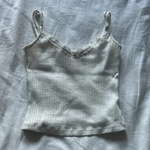 Vit linne med spets kanter  - Supersöt vit linnetopp med smala axelband och v-ringning dekorerad med spets. Toppen är ribbad och har en figurnära passform som känns både enkel och trendig. Perfekt att styla med jeans eller kjol för en fräsch look. Barn storlek och går rätt högt upp. Magtröja!!! 