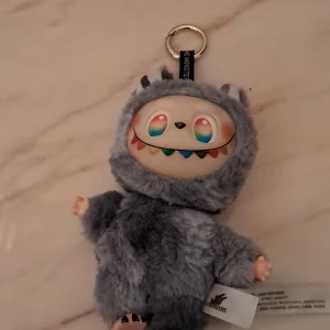 Grå plysch monster-nyckelring - Söt och fluffig nyckelring från The Monsters i form av ett grått monster med färgglada detaljer i ansiktet. Perfekt att hänga på väskan eller använda som accessoar. Mjuk pälsliknande yta och metallring för enkel fastsättning.